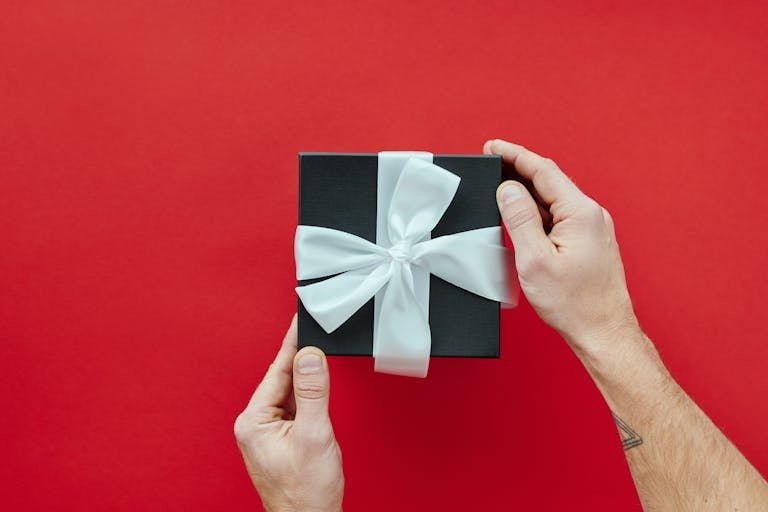 Elegant black gift box with a white ribbon held by hands against a vibrant red background.