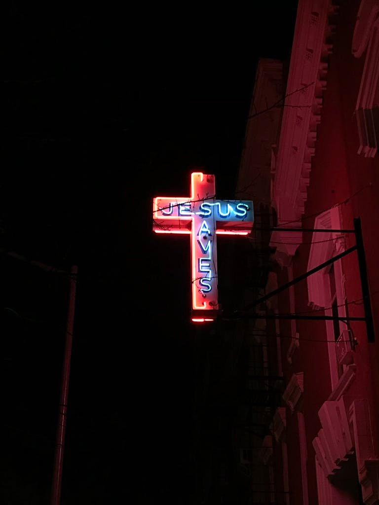 A glowing neon cross sign that reads 'Jesus Saves' against a dark night sky.