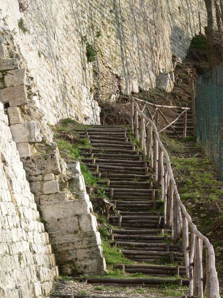 Rustic stone steps with wooden railings ascend a hill alongside a stone wall, evoking a sense of history.