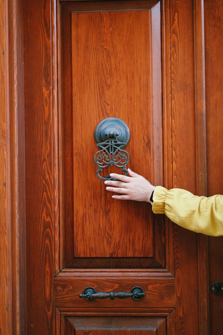 A person in a yellow sleeve reaching for a classic door knocker on a wooden door.