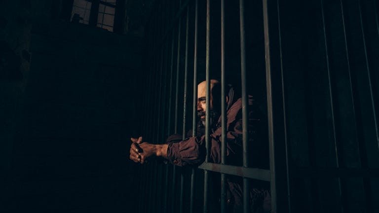 A solitary man stands locked behind bars in a dark prison setting.