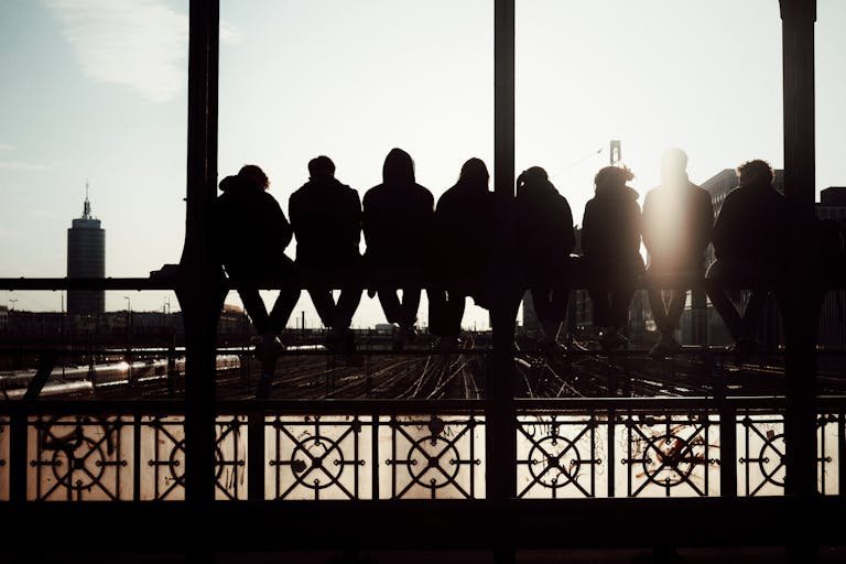 Group of seven people sitting on a bridge railing, silhouetted against a sunset sky in an urban setting.
