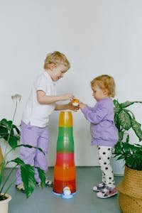 Two children enjoy playful learning with colorful stacking cups indoors surrounded by plants.